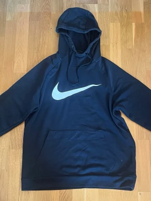 Svart Nike hoodie med stor logga - Snygg svart hoodie från Nike med stor vit Swoosh-logga på bröstet. Klassisk känguruficka framtill och justerbar huva med dragsko. Tillverkad i mjukt och bekvämt material, perfekt för en avslappnad streetwear-look.