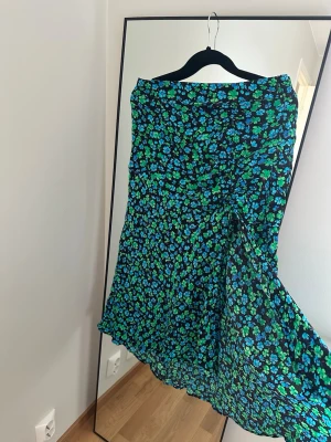 Blommig långkjol i blått och grönt - Säljer en flowy långkjol med blommigt mönster i blått och grönt på svart botten. Kjolen har en lätt volangkant och omlottkänsla, perfekt för dig som gillar färg och mönster. Superskön och snygg till sneakers eller sandaler.