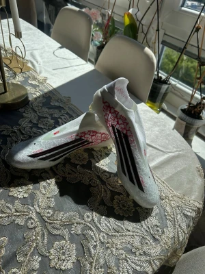 Vita Adidas fotbollsskor med mönster - Säljer ett par vita Adidas fotbollsskor med svarta ränder och röda detaljer. Skorna har ett unikt mönster över hela ovandelen och är tillverkade i syntetmaterial. Designen är modern och strömlinjeformad, perfekta för dig som vill sticka ut på planen. Denna sko är lamin yamal edition, aldrig anvönda söljs pågrund av att de är för små på mig fick de som present