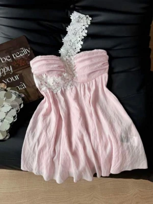 Ljusrosa oneshoulder klänning med spets - Supersöt ljusrosa kortklänning med unik oneshoulder-design och vit spetsdetalj över axeln. Klänningen har en lätt rynkad byst och ett flowy fall. Perfekt för dig som vill sticka ut med en romantisk look.