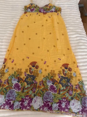 Gul långklänning med blommigt mönster - Säljer en somrig gul långklänning med tunna axelband och färgglatt blommigt mönster i lila, vitt och grönt längs nederkanten. Klänningen har en smockad överdel och är tillverkad i ett lätt och luftigt material, perfekt för varma dagar.