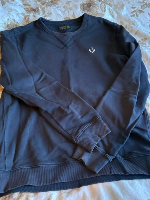 Mörkblå sweatshirt med broderad logga - En stilren mörkblå sweatshirt med rund hals och ribbade muddar. Tröjan har en liten broderad logga på bröstet och är tillverkad i mjukt bomullsmaterial. Perfekt för en avslappnad look och enkel att matcha med jeans eller byxor.