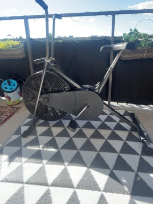 Träningscykel med fläkt och display - Säljer en svart och silver träningscykel med stor fläkt framtill och digital display. Cykeln har ett brett, bekvämt sadel och två handtag som rör sig när du trampar. Perfekt för hemmaträning och kondition. Robust metallram och plastdetaljer.