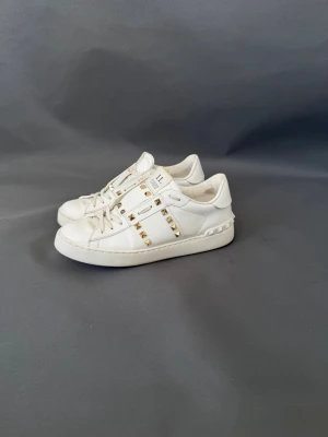 Vita sneakers med gulddetaljer från Valentino - Säljer ett par vita sneakers från Valentino med guldfärgade nitar längs sidorna och klassisk snörning. Skorna har en stilren design med exklusiv känsla och är tillverkade i skinn. Perfekta för dig som vill ha något extra till din outfit.