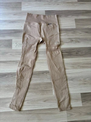 Beige leggings från Stronger - Snygga beige leggings från Stronger med hög midja och ribbad struktur upptill. Tillverkade i ett stretchigt material av 94% polyamid och 6% elastan för en skön och följsam passform. Perfekta för både träning och chill.
