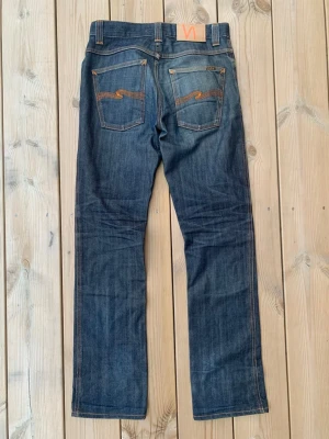 Nudie Jeans Low Slim Jim blå jeans - Säljer ett par klassiska blå jeans från Nudie Jeans, modellen Low Slim Jim. Jeansen har en slim passform, snygga kontrastsömmar och ikoniska bakfickor med orange brodyr. Tillverkade i 100% bomull och har en cool tvättad look.