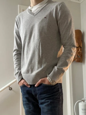 Ljusgrå V-ringad tröja Tommy Hilfiger - Snygg ljusgrå V-ringad tröja från Tommy Hilfiger med diskret logga på bröstet. Tröjan har långa ärmar och ribbade muddar vid ärmslut och nederkant. Perfekt att bära över en skjorta eller t-shirt för en clean och stilren look.