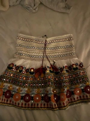 Broderad kortkjol från Zara - Färgglad kortkjol från Zara med broderade blommor, speglar och snörning i midjan. Kjolen har vita och röda detaljer, inslag av blått, grönt och orange samt volangkant. Perfekt för dig som gillar boho-stil och unika detaljer.
