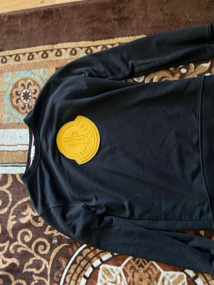 Svart sweatshirt från Moncler - Säljer en svart sweatshirt från Moncler med en stor gul Moncler-logga på bröstet. Tröjan har rund halsringning och långa ärmar. Perfekt för dig som gillar streetwear och vill ha något som sticker ut.