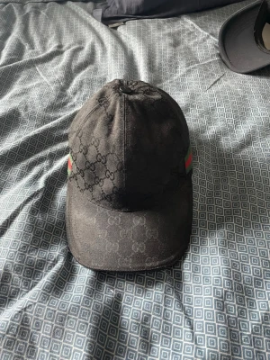 Svart Gucci keps med GG-mönster - Snygg svart keps från Gucci med klassiskt GG-monogram över hela kepsen. På sidorna finns de ikoniska gröna och röda ränderna. Justerbar rem bak för perfekt passform. Tillverkad i slitstarkt tyg som känns lyxigt och trendigt.