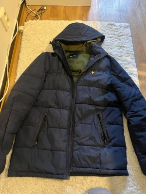 Marinblå pufferjacka från Lyle & Scott - Snygg marinblå pufferjacka från Lyle & Scott med huva och dragkedja. Jackan har två framfickor med dragkedja och en olivgrön insida. Dragkedjan behöver bytas det är därför den är så billig 