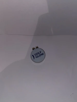 Golf Glow markör  - Säljer en rund markeringsknapp från Golf Glow i metall med magnetfäste. Framsidan är vit med blå text och en siluett av en golfspelare. Perfekt att ha på kepsen eller bältet när du spelar golf.