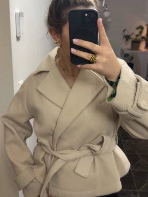 Beige kappa med knytskärp - Stilren beige kappa med bred krage och knytskärp i midjan. Kappan har dekorativa knappar på ärmsluten och en mjuk, ull-liknande yta. Perfekt för dig som vill ha en klassisk och trendig look till hösten eller vintern.