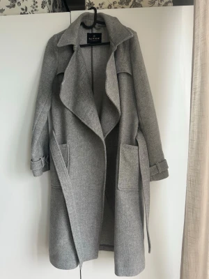 Kappa - Snygg grå kappa från Review med oversized krage, stora framfickor och bälte i midjan. Kappan har ett mjukt, ull-liknande material och justerbara band vid ärmsluten. Perfekt för kyliga dagar och ger en stilren look.