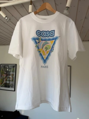 Vit Casablanca Paris t-shirt med tryck - Säljer en vit t-shirt från Casablanca Paris med färgglatt grafiskt tryck framtill i blått, gult och grönt. Klassisk passform med rund hals och korta ärmar. Perfekt för dig som gillar streetwear och vill sticka ut lite extra.