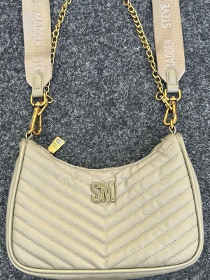 Beige quiltad handväska Steve Madden - Trendig beige handväska från Steve Madden med quiltat mönster och guldfärgade detaljer. Väskan har en bred axelrem med kedja och textlogga, samt ett SM-emblem framtill. Perfekt accessoar för att lyfta din outfit.