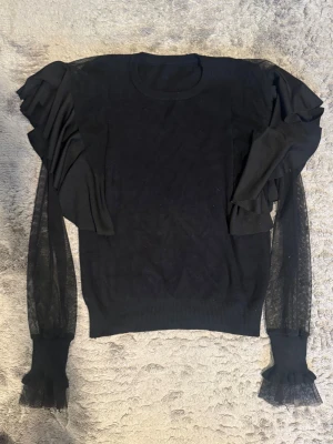 Svart stickad tröja med volangärmar - Hittade den trendig svart stickad tröja med volangdetaljer på axlarna och transparenta meshärmar i mammas garderob och kände att den behöver ett nytt hem💞. Den har rund halsringning och ribbade muddar med volangavslut. Tröjan är använd få gånger! Den är super snygg och passar för både vanlig dag men även en fest💕! 