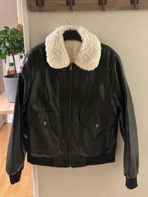 Svart skinnjacka med teddykrage storlek L - Snygg svart skinn imitation med vit teddykrage och dragkedja framtill. Två stora fickor med lock och knapp på framsidan. Jackan har ribbade muddar vid ärmslut och nederkant för extra komfort. Perfekt för dig som vill ha en cool och tidlös look. 