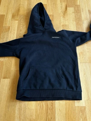 Svart hoodie från Peak Performance - Klassisk svart hoodie från Peak Performance med broderad logga på bröstet. Hoodien har stor huva och en rymlig magficka framtill. Tillverkad i mjukt material och har ribbade muddar vid ärmslut och nederkant. Perfekt för en avslappnad och stilren look.
