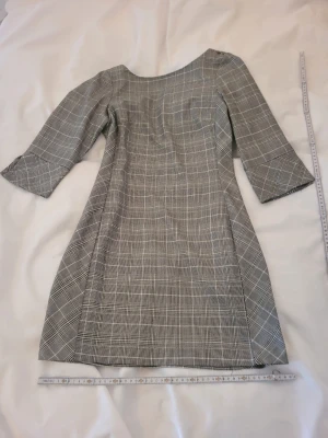 Grå rutig klänning med trekvartsärm - Snygg grå klänning med rutigt mönster och trekvartsärm. Klänningen har en lätt utsvängd form och rund halsringning. Perfekt för dig som gillar klassisk stil med modern twist.