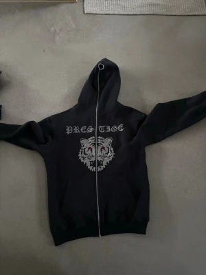 Svart hoodie med tigertryck - Svart hoodie med dragkedja ifrån aimforthemoon andvänd cirka 2-3 gångerfrån. På bröstet finns ett stort tryck av en tiger med röda ögon och texten 'PRESTIGE' ovanför. Hoodien har huva med dragsko och två fickor framtill. Perfekt för en streetwear-look.
