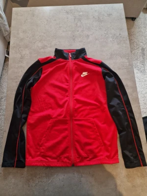 Röd och svart Nike zip up - Sportig zip-jacka från Nike i rött med svarta ärmar och röda detaljer. Jackan har hög krage, två fickor fram och vit Nike-logga på bröstet. Tillverkad i mjukt och bekvämt material, perfekt för träning eller chill.