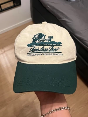 Aime Leon Dore beige och grön keps - Snygg keps från Aime Leon Dore med beige krona och mörkgrön skärm. Framtill finns ett broderat motiv av Unisphere och texten 'Aime Leon Dore' samt 'Queens, New York, The World's Borough'. Justerbar baktill och tillverkad i bomull.