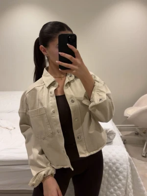 Beige cropped jeansjacka - Snygg beige jeansjacka i croppad modell med avslappnad passform. Jackan har stora bröstfickor, klassisk krage och silverfärgade knappar framtill. Perfekt att slänga över en topp för en trendig look.