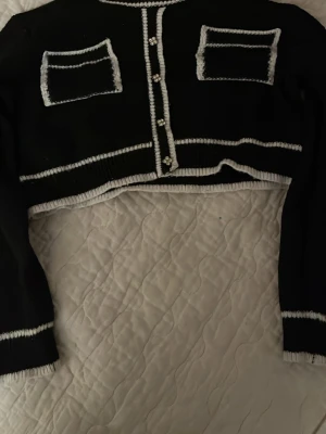 Svart cropped kofta från Knit Mix - Stilren svart cropped kofta från Knit Mix med vita kontrastsömmar och dekorativa pärlknappar framtill. Två fejkade bröstfickor och ribbade kanter ger en trendig look. Perfekt att styla över en topp eller klänning.