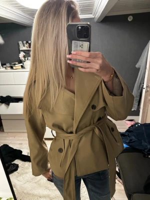 Kort beige trenchcoat med knytskärp - Snygg kort trenchcoat i beige med dubbelknäppning och brett knytskärp i midjan. Klassisk krage och stora knappar framtill. Jackan har en avslappnad passform och är tillverkad i ett slätt tyg som ger en stilren look.