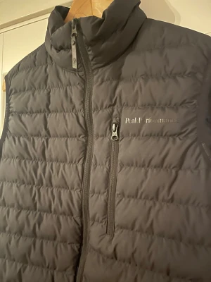 Svart dunväst Peak Performance  - Stilren svart Peak Performance dunväst med hög krage och quiltad design. Västen har dragkedja framtill, två sidofickor med dragkedja och en bröstficka med dragkedja. 