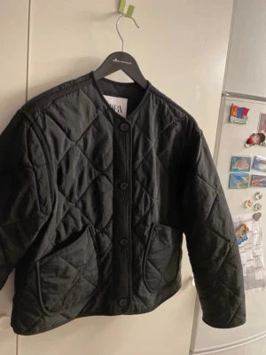 Svart quiltad vindjacka från Zara - Stilren svart vindjacka från Zara med quiltat mönster och stora fickor framtill. Jackan har rund halsringning, knäppning med stora knappar och en avslappnad passform. Perfekt för lager-på-lager och enkel att matcha till olika outfits.