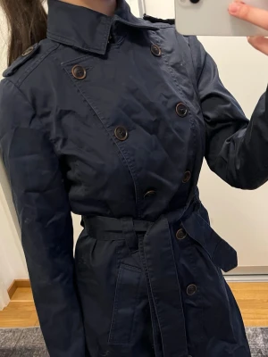 Mörkblå trenchcoat från Morris - Stilren mörkblå trenchcoat från Morris med dubbelknäppning, axelklaffar och avtagbart midjebälte. Klassisk krage och dekorativa knappar i brunt. Lätt figursydd passform och slät yta i ett tunt material, perfekt för vår och höst.