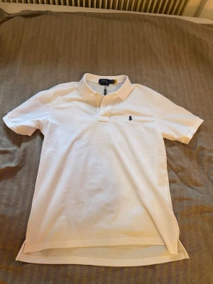 Vit pikétröja från Polo Ralph Lauren - Klassisk vit pikétröja från Polo Ralph Lauren i bomull med korta ärmar och blå broderad logga på bröstet. Tröjan har krage och knappar framtill samt en slim fit passform.  Storlek M herr
