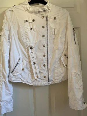Vit vindjacka med detaljer - Snygg vit vindjacka med flera silverfärgade knappar och dragkedjor, hög krage och lång ärm. Jackan har coola detaljer som fickor med dragkedja och dekorativa sömmar. Perfekt för dig som gillar en sportig och modern stil.
