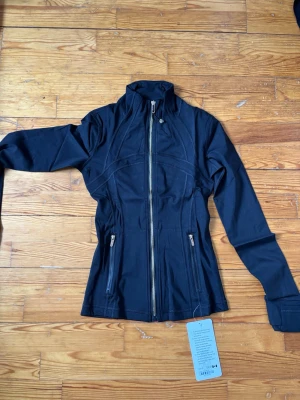 Svart träningsjacka från Lululemon - Sportig svart vindjacka från Lululemon med hög krage och figurnära passform. Jackan har dragkedja framtill, två fickor med dragkedja och långa ärmar. Perfekt för aktiva dagar och snygg till träningslooken.