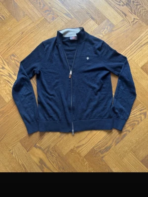 Mörkblå ziptröja från Morris - Mörkblå ziptröja från Morris med diskret logga på bröstet. Tröjan har hög krage, hel dragkedja och ribbade muddar. 100 merino ull 