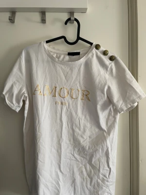 Vit t-shirt med guldknappar och text - Vit t-shirt med guldfärgad text 'AMOUR PARIS' på bröstet och tre dekorativa guldknappar på ena axeln. Klassisk rund halsringning och korta ärmar. Perfekt för en trendig och enkel look.