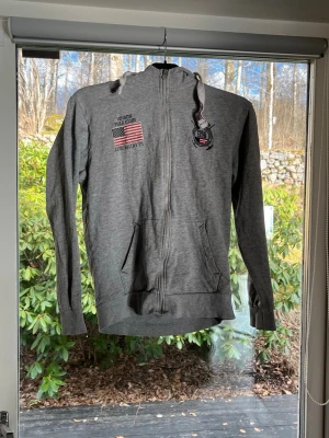 Grå zip hoodie Vision Polo Club - Grå hoodie med dragkedja från Vision Polo Club. Har två fickor fram, broderade märken och amerikansk flagga på bröstet. Justerbar huva med snören och ribbade muddar vid ärmslut och nederkant. Perfekt för en avslappnad streetwear-look. Hör av er vid frågor eller prisförslag!