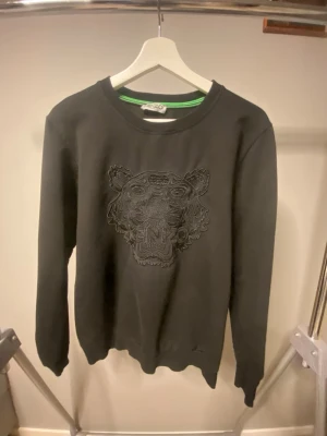 Svart sweatshirt från Kenzo med tiger - Säljer en svart sweatshirt från Kenzo med det ikoniska tigermotivet broderat på bröstet. Tröjan har rund halsringning, långa ärmar och ribbade muddar. Tillverkad i mjukt bomullsmaterial och har en clean, stilren look med diskreta detaljer.