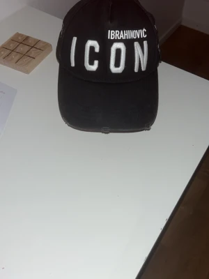 Svart DSQUARED2 ICON Ibra keps - Säljer en svart keps från DSQUARED2 med stor vit brodyrtext 'ICON' och 'IBRAHIMOVIC' framtill. På sidan står DSQUARED2 och detaljer om ett samarbete. 