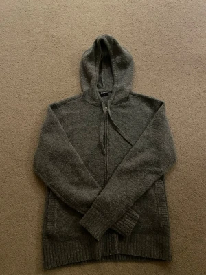 Mörkgrå hoodie med dragkedja - Mysig mörkgrå hoodie med dragkedja framtill och huva. Tillverkad i ett mjukt, stickat material som känns skönt mot huden. Långa ärmar och ribbade muddar vid ärmslut och nederkant ger en avslappnad look. Perfekt för chill dagar eller när du vill vara extra bekväm.