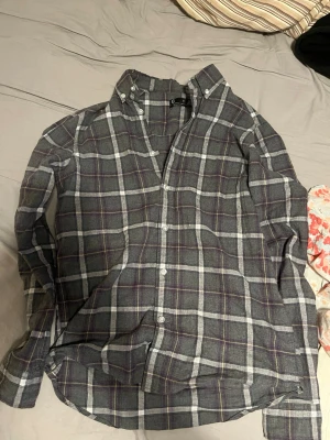 Grå rutig skjorta från Cubus  - Klassisk grå rutig skjorta från Cubus med vita och lila ränder. Skjortan har button-down krage, långa ärmar och vita knappar framtill. Perfekt för en avslappnad men ändå stilren look.