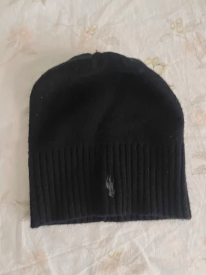 Svart mössa  - Svart ribbstickad mössa från Polo Ralph Lauren med klassisk broderad logga framtill. Mössan har en enkel och stilren design, perfekt för kalla dagar. Materialet känns mjukt och bekvämt mot huden.