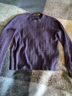 Lila kabelstickad tröja från Polo Ralph Lauren - Snygg lila kabelstickad tröja från Polo Ralph Lauren med klassisk röd logga broderad på bröstet. Tröjan har rund halsringning och långa ärmar, perfekt för lager på lager och en stilren look.
