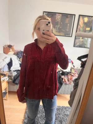 Vinröd skjorta - Säljer en vinröd skjorta med långa ärmar och knappar framtill. Skjortan har två bröstfickor med lock och en loose passform som ger en avslappnad look. Perfekt att styla med jeans eller över en t-shirt.