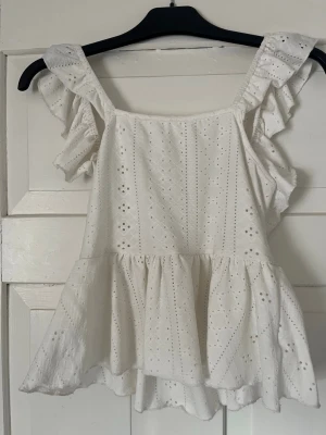 Vit volangblus med broderade detaljer - Supersöt vit blus med volangärmar och broderade hålmönster. Toppen har en luftig passform, omlottdetalj och knyt i ryggen. Perfekt för sommarens alla tillfällen och ger en romantisk vibe.