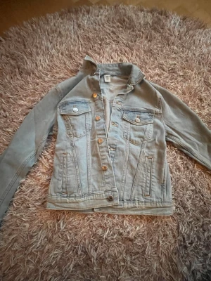 Ljusblå klassisk jeansjacka - Säljer en ljusblå jeansjacka med klassisk passform och silverfärgade knappar. Jackan har två bröstfickor med lock och knapp samt två sidofickor. Perfekt att slänga över en hoodie eller t-shirt för en avslappnad look.