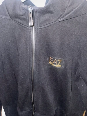 Svart hoodie från EA7 Emporio Armani - Svart hoodie med dragkedja från EA7 Emporio Armani. Klassisk design med huva och diskret logga i guld och svart på bröstet. Tillverkad i mjukt material som känns skönt mot huden. Perfekt för en avslappnad och stilren look. Betalning genom vanlig swish! 