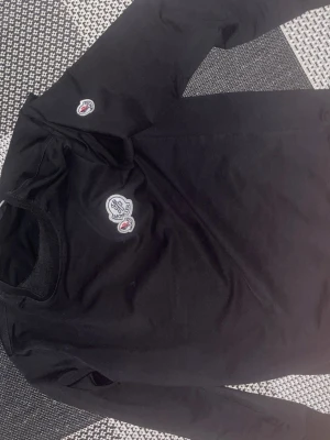 Svart långärmad topp från Moncler - Snygg svart långärmad topp från Moncler med rund halsringning och ikoniska Moncler-loggor på bröstet och ärmen. Tillverkad i mjuk bomull som känns skön mot huden. Perfekt för dig som gillar stilrena och exklusiva plagg.